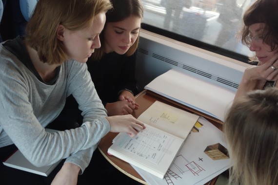 Vier junge Frauen beugen sich über Tisch und betrachten die drauf liegenden Unterlagen (Gestaltungskonzept)