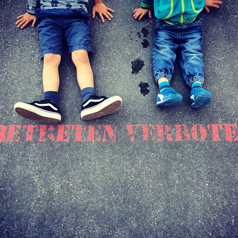 Kinder sitzend auf Asphalt. Vor Füßen steht auf dem Asphalt in roter Schrift: Betreten verboten!