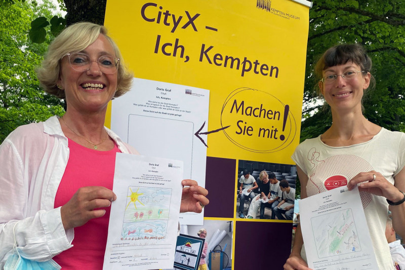 Zwei Frauen stehen vor gelbem Banner mit Schriftzug &quot;CityX – Ich, Kempten. Machen Sie mit!&quot; und zeigen Buntstiftzeichnungen und stehen vor 