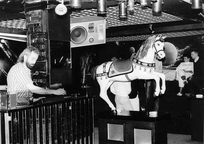 Schwarz-Weiß-Bild: Mann an DJ-Pult in Dikothek. Nebem ihm Holzfigur Pegasus.