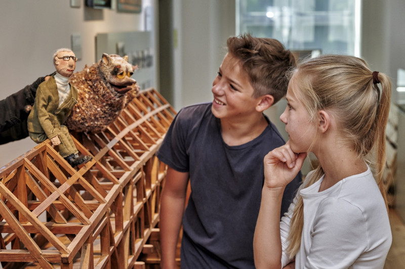 M&auml;dchen und Junge an Br&uuml;ckenmodell, auf dem Handpuppen Uhu und Mann sitzen