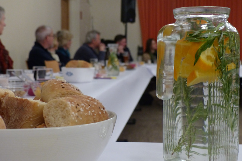 Wasserkaraffe und Schale mit Baguette auf einem Tisch, im Hintergrund Menschen im Gespräch
