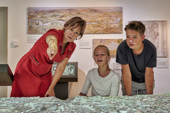 eine Frau in rotem Kleid erkl&auml;rt einem M&auml;dchen und einem Jungen ein Stadtmodell