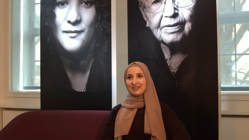 eine Frau mit Kopftuch sitzt vor zwei schwarz-weiß Fotos mit Porträtbildern von Frauen