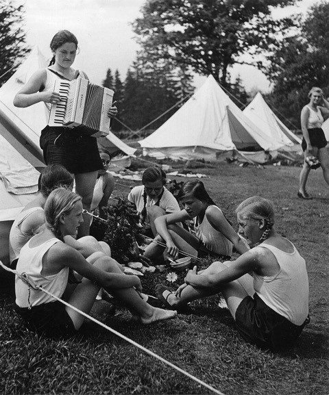 schwarz wei&szlig; Foto mit sitzenden M&auml;dchen in einem Zeltlager, eines spielt ein Akkordeon 