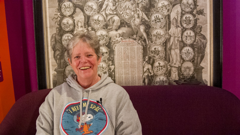 eine lachende Frau mit Kurzhaarschnitt und Kapuzenpulli sitzt auf einem Sofa im Museum und erz&auml;hlt